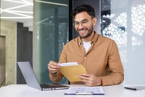 Happy indian man sitting at desk in modern office and smilingly opening and reading received letter. photo