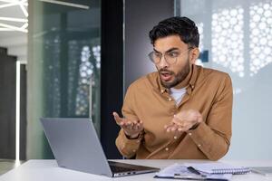 A confused man wearing glasses gestures at his laptop screen while working in a modern office, expressing frustration or misunderstanding. photo