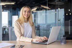 Portrait of a young businesswoman sitting in the office at the desk, working on a laptop, smiling and looking at the camera. photo