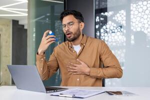 Asthmatic man uses inhaler for relief while working on a laptop in a modern office environment. photo