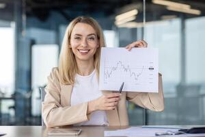 Portrait of a young business woman sitting in the office and showing documents with graphs to the camera with a pen. Presents the steady growth of the company's income, talks on a call. photo