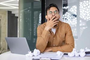 Tired businessman yawning at his desk surrounded by a laptop and crumpled papers in a modern office setting. Concept of fatigue, overworked, and stress. photo