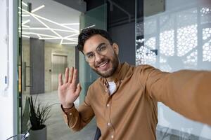 Smiling man wearing glasses and waving at camera in a modern office setting during a call. Friendly and approachable atmosphere. photo