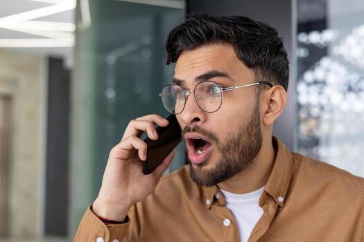 de cerca de un hombre demostración conmoción y sorpresa mientras Hablando en un teléfono llamada en un interior ajuste. foto