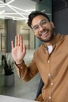 A happy, smiling man waves during a call in a modern office. The image captures friendly interaction and professional communication. photo