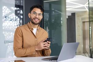 Smiling young professional using smartphone while sitting at desk with laptop in modern office environment. Business technology and communication concept. photo
