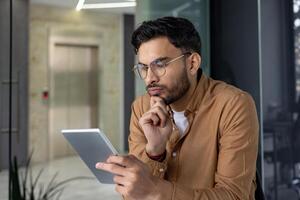Man in casual clothing thoughtfully using a digital tablet in a modern office setting. Person engaging in work or study. photo