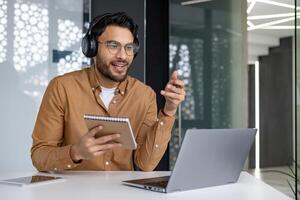 Man wearing headphones attending an online webinar while taking notes. He is sitting at a modern office desk with a laptop and notebook. photo