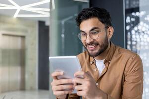 Young Hindu programmer close up inside office using application on tablet computer, male developer testing new software, businessman at workplace wearing shirt and glasses. photo