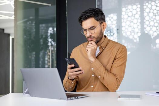 un enfocado empresario comprobación su teléfono mientras trabajando en un ordenador portátil en un moderno oficina configuración, retratar concentración y productividad. foto