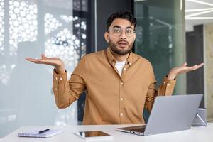 Young man with glasses in an office environment sitting in front of a laptop, expressing confusion and uncertainty with open palms and shrugging gesture. photo