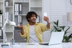 Young man in a modern office taking a selfie with his smartphone, expressing joy and confidence while working at his desk with a laptop. photo