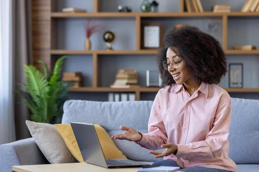 un mujer con Rizado pelo es sonriente en un sofá como ella trabajos en su computadora portátil, rodeado por estantería con libros y decoración. esta acogedor y productivo hogar oficina ofertas un cómodo espacio de trabajo foto