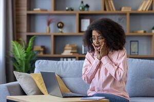 Shocked woman sitting on a couch at home, using a laptop and expressing astonishment. Surprised reaction in a domestic setting. photo