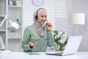 Smiling woman in a hijab using a headset on a call, working on a laptop in a modern home office. photo