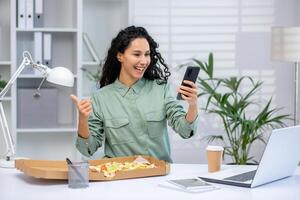 Cheerful woman having lunch at her desk while calling with smartphone, showing thumbs up in a modern office setup with laptop and coffee. photo