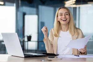 A professional woman in a modern office setting celebrating a business achievement with a fist pump and holding documents. photo