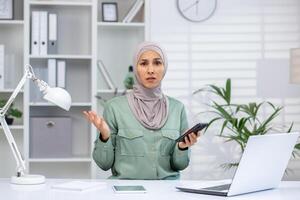 Woman in hijab working from home office, holding smartphone and gesturing in confusion. Modern workspace with laptop and documents. photo