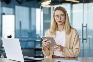 Focused young professional using a tablet while sitting in a well-lit office space with a laptop nearby. This image captures the essence of modern workplace flexibility and technology integration. photo