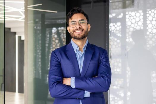 retrato de joven exitoso indio hombre dentro oficina, hombre sonriente y mirando a cámara con cruzado brazos, satisfecho empresario trabajador en negocio traje, jefe financiero. foto