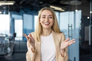 A young professional woman talks enthusiastically, gesturing with her hands in a modern, well-lit office setting. She looks directly at the camera, her expression joyful and confident. photo