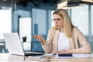 A young business woman is working in the office on a laptop, sitting at the table and talking on a call. Upset, she holds a meeting, discusses financial problems. photo