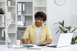 Young professional uses laptop, documents, and calculator while working from home in a modern, well-organized office. photo