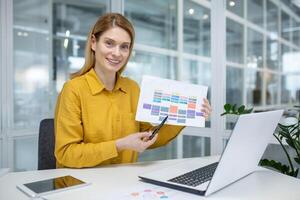 Confident woman in yellow shirt showing a printed schedule while sitting at her desk in a bright modern office with a laptop. photo