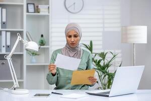 Businesswoman in hijab reading a document with a worried expression at her office desk, laptop open, files and lamp nearby. photo