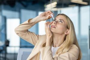A businesswoman in a beige suit applies eye drops while looking upward, expressing discomfort, in a bright office environment. photo