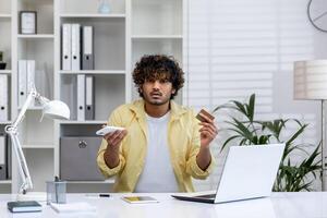 Confused man holding a credit card and smartphone while sitting at an office desk with a laptop, displaying uncertainty. photo