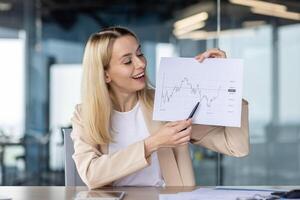 A smiling young business woman holds in her hands and shows to the camera documents with work results, graphs, a project. Satisfied with the company's financial success. photo