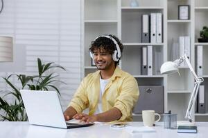 Man wearing headphones and working on a laptop in a comfortable, well-organized home office setting, emphasizing remote work and technology. photo