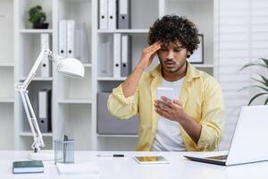 A stressed man holds his smartphone while working in an office. He appears frustrated and concerned about a business issue. photo
