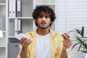 A confused man holding a mobile phone and credit card, looking perplexed while standing in an office environment with shelves and files in the background. photo