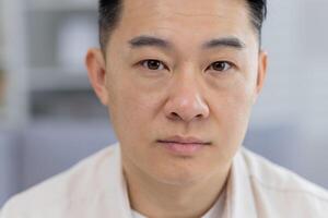 A detailed close-up portrays a middle-aged Asian man with a contemplative, serious expression, captured indoors with a soft focus background. photo