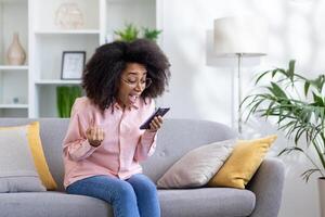 Young beautiful woman sitting on sofa in living room with phone in hands, winner happy with good news received on smartphone, african american woman happy and celebrating achievement results. photo