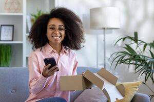 en el cómodo vivo habitación, un mujer felizmente desempaqueta un entrega en un gris sofá, participación su teléfono, rodeado por plantas, con un grande sonrisa, cultivando un de moda, contemporáneo hogar ajuste foto