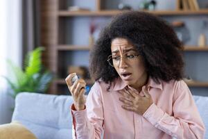 In the living room, a woman with curly hair and glasses sits on a couch, looking concerned, holding an inhaler. Plants and shelves fill the background providing an indoor scene of comfort and care photo