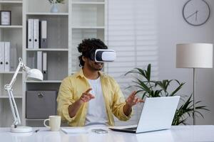 Man wearing VR headset and interacting with virtual environment at his office desk, with a laptop in front of him. photo