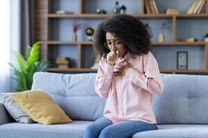 A middle-aged woman with glasses is coughing into her hand while sitting on a gray couch. The background features shelves with books and plants, creating a cozy living room atmosphere. photo