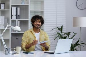 A cheerful man participates in an online course, taking notes while watching a webinar on a laptop in a modern home office. photo