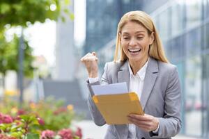 A joyful businesswoman, professionally dressed, celebrates outdoors in an office courtyard upon receiving positive news in an envelope. She reflects happiness and success in her expression photo