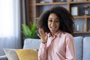 In a welllit cozy living room, a cheerful woman with glasses is waving her hand. The room is furnished with shelves, plants, and a comfy sofa adorned with yellow and gray pillows photo
