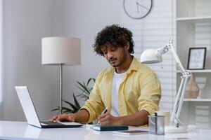 Man in a yellow shirt working remotely from home, using a laptop and taking notes in a notepad at a modern desk. photo