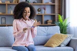 un mujer en el vivo habitación reacciona felizmente a un positivo el embarazo prueba, sentado cómodamente en un sofá, expresando alegría con un sonrisa. el moderno interior agrega a el acogedor atmósfera foto