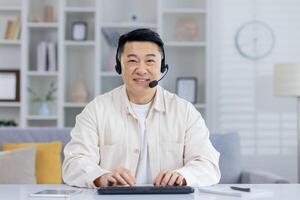 A middle-aged Asian man smiles warmly while using a headset and working on a keyboard in a bright, modern home office setting. photo