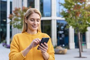 A cheerful woman in a yellow sweater happily using her smartphone while standing in a modern cityscape with trees and office buildings in the background. photo