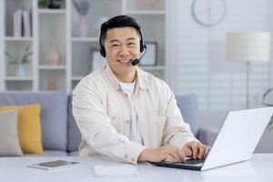 Happy middle-aged Asian man using a headset and laptop in a well-decorated home office. He exudes professionalism and contentment while working remotely. photo