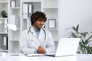 Confident ethnic doctor providing virtual healthcare services in a modern clinic setting, using a headset and laptop. photo